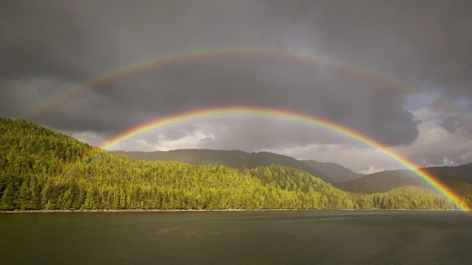A vibrant double rainbow arches across a dramatic sky over a densely forested coastline.