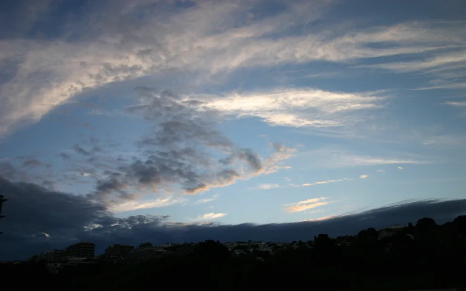 A wide shot of a city skyline silhouetted against a dramatic sky filled with wispy clouds at dusk.