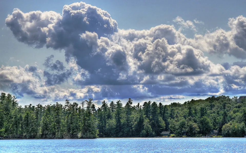 Dramatic, puffy white and grey clouds fill the sky above a dense green forest and a blue lake.