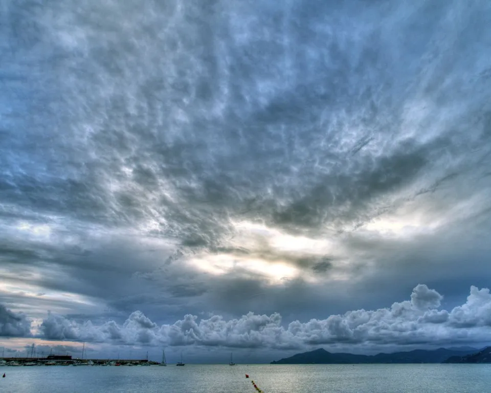 A vast expanse of dramatic, textured clouds fills the sky above a calm sea with a distant, dark mountain range.