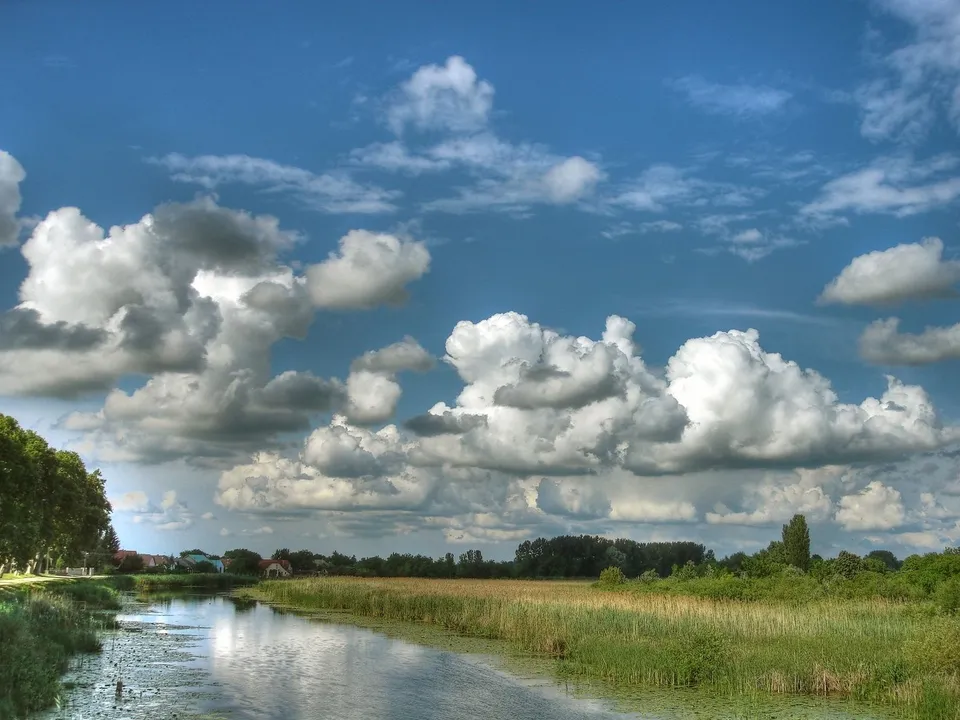 A wide shot of a calm river reflecting a bright blue sky filled with large, fluffy cumulus clouds.