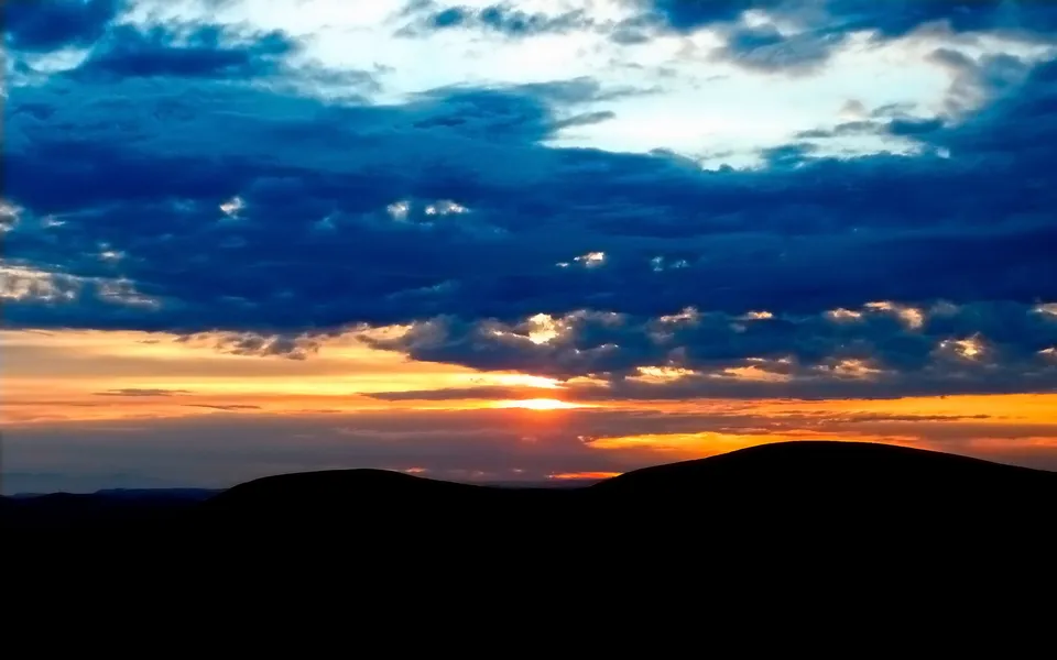 A vibrant sunset with dark blue clouds and orange hues over silhouetted desert hills.