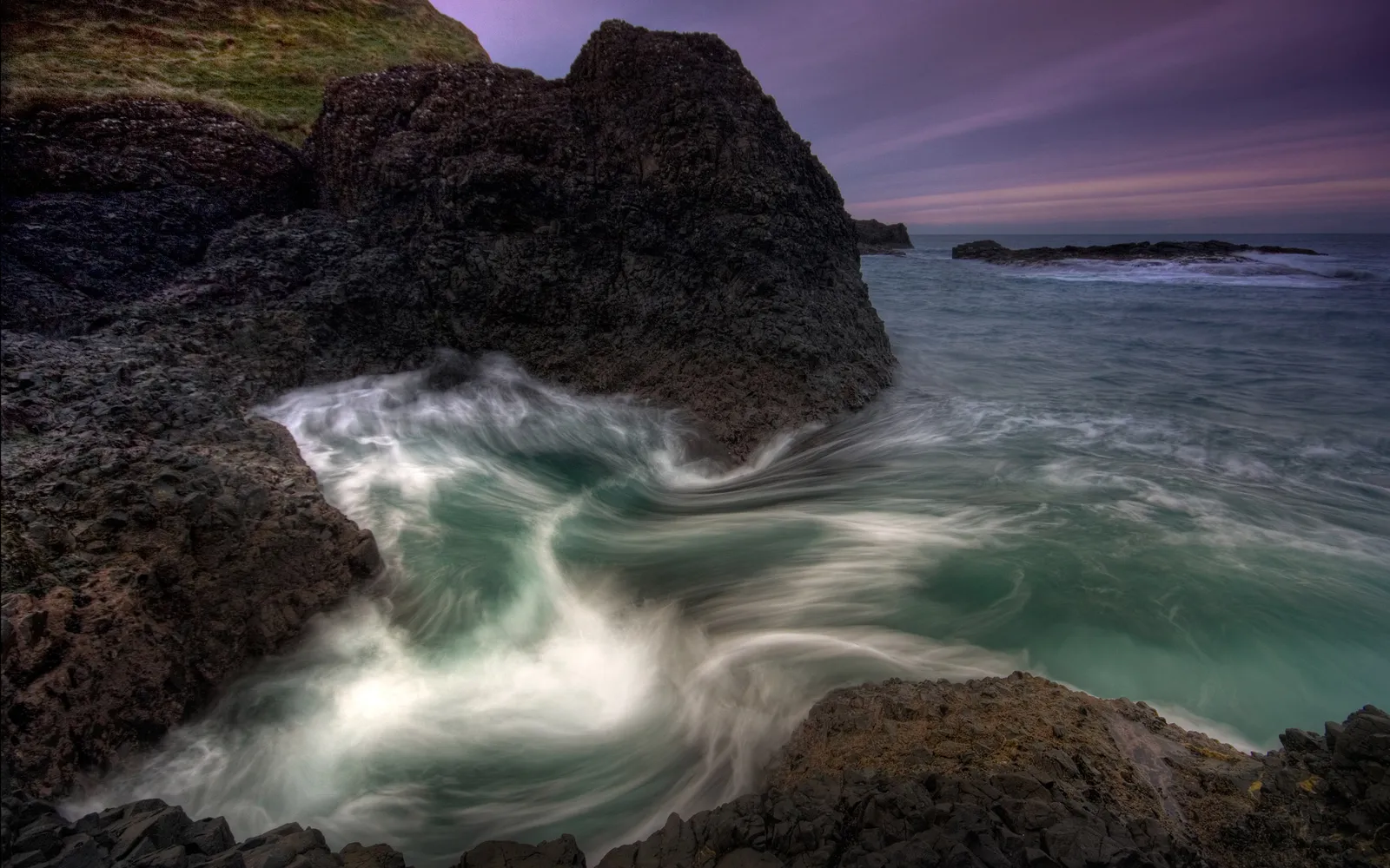 Waves crash against dark volcanic rocks at dusk with a purple sky.