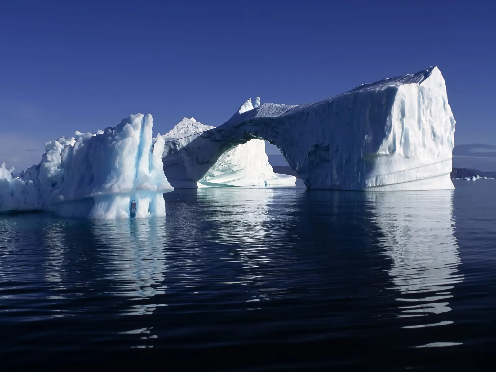 A striking natural arch formed within a massive iceberg, reflecting on still, dark water under a bright blue sky.