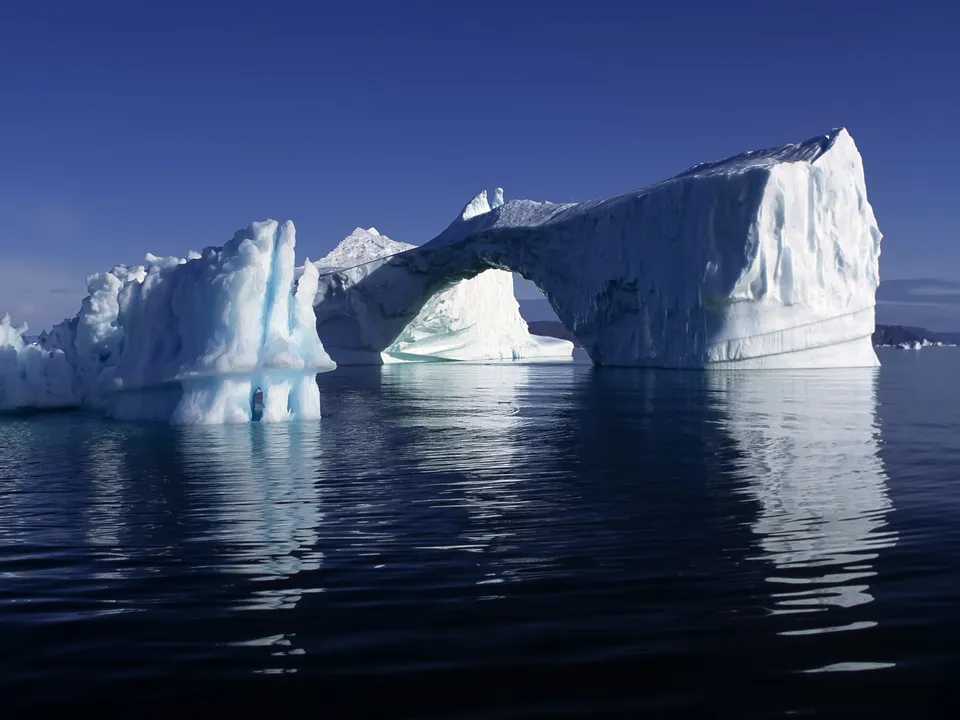 A striking natural arch formed within a massive iceberg, reflecting on still, dark water under a bright blue sky.