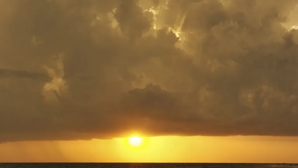 The sun sets over the ocean, casting golden light through dramatic clouds with visible sun rays.