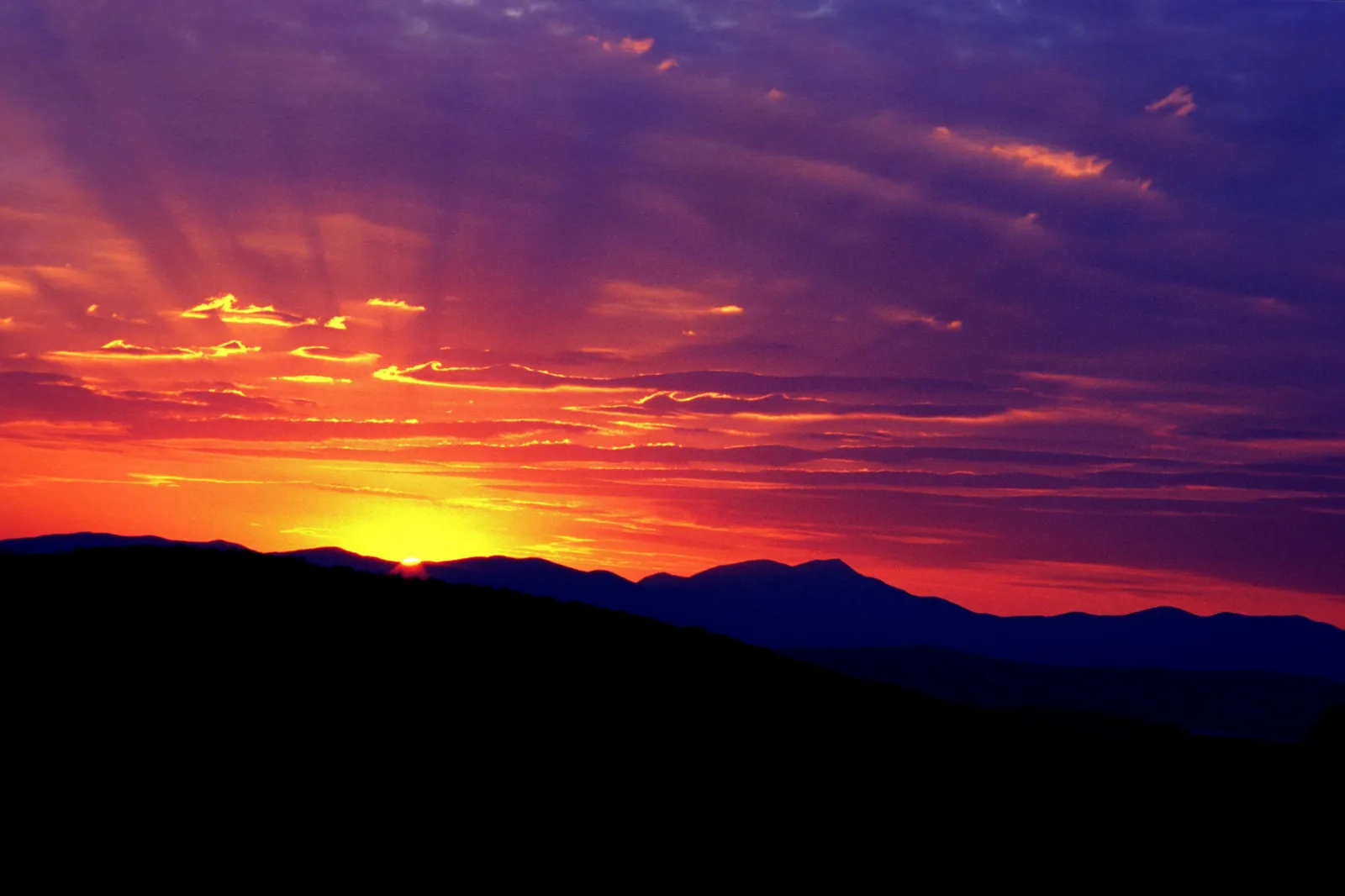 Dramatic sun rays pierce through clouds during a vibrant sunrise over silhouetted mountain peaks.