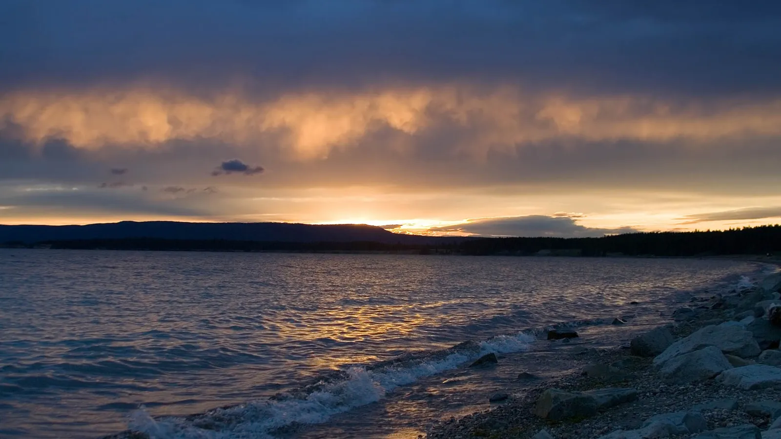 A dramatic sunset casts a warm glow on clouds over distant mountains, with gentle waves on a rocky shore.