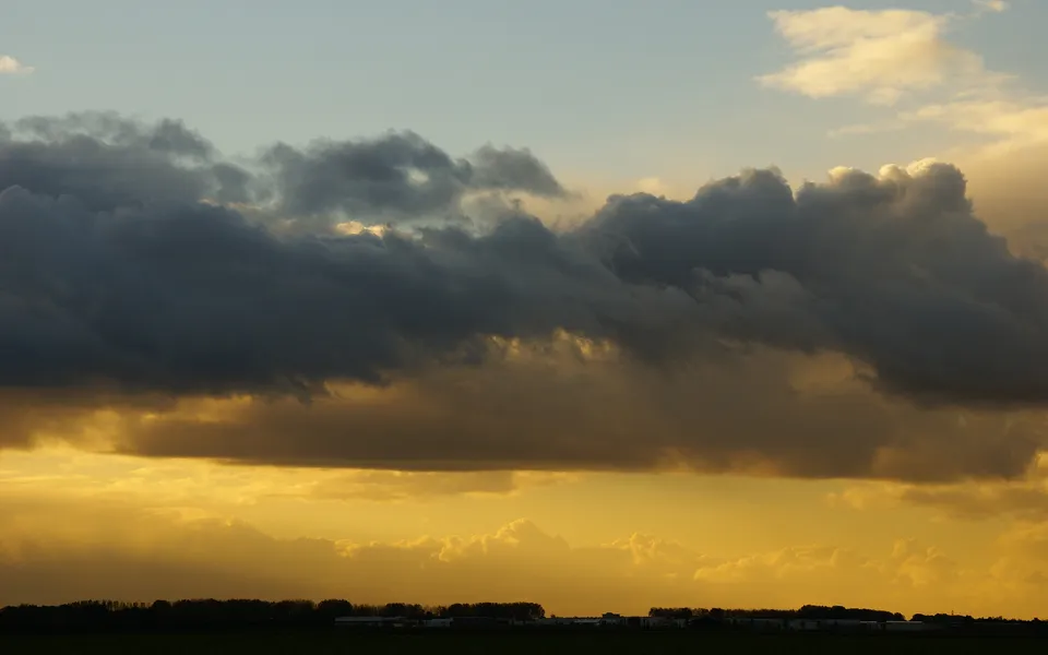 Dark, heavy clouds are illuminated by the golden light of a sunset over a distant treeline and fields.