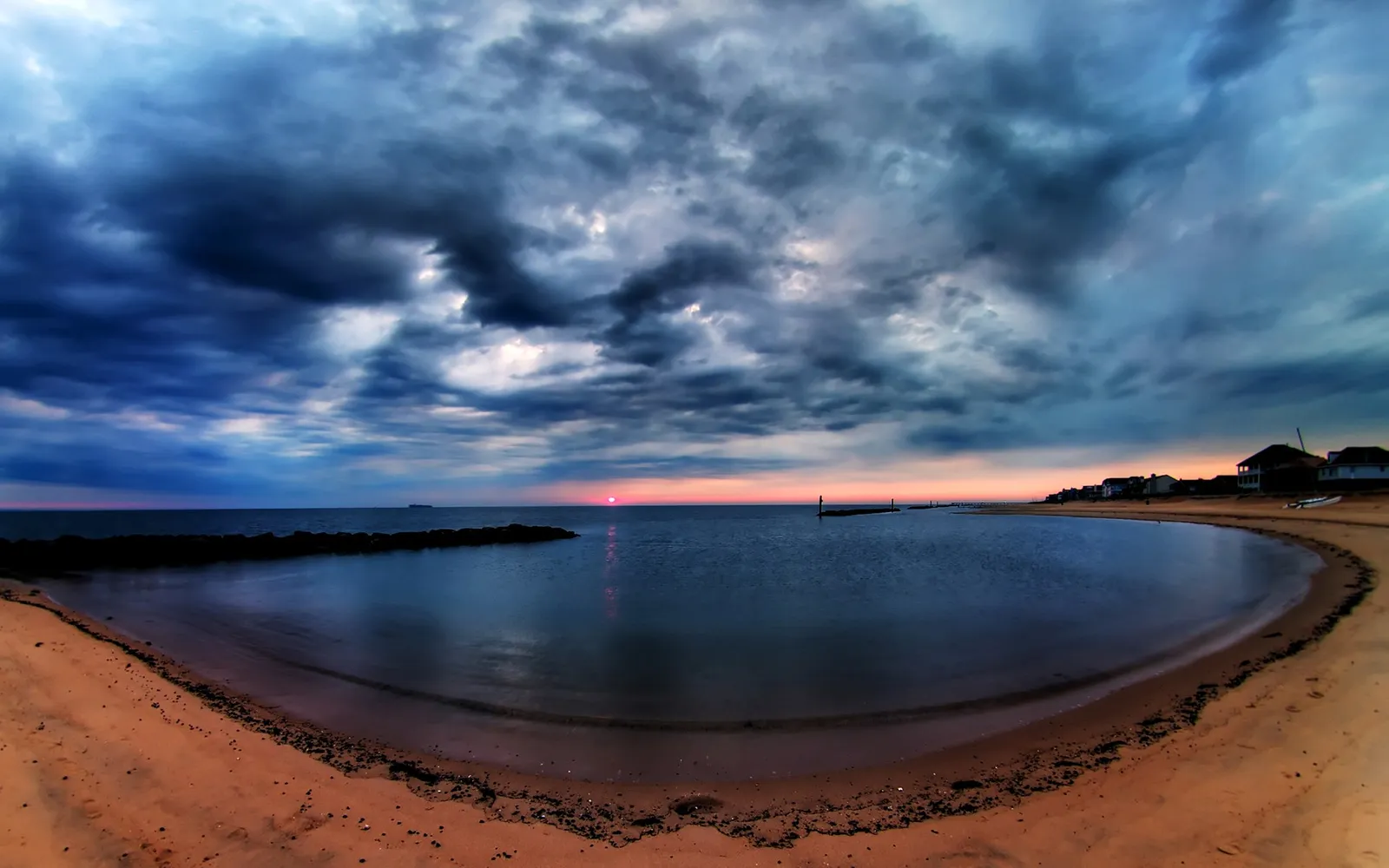 A dramatic sunset with dark clouds over a calm ocean, viewed from a sandy beach.