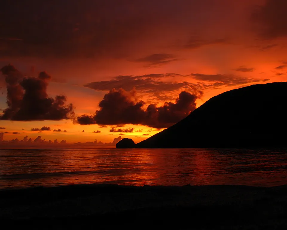 A vibrant orange and red sunset over the ocean, with dark clouds and a silhouetted island and hill.