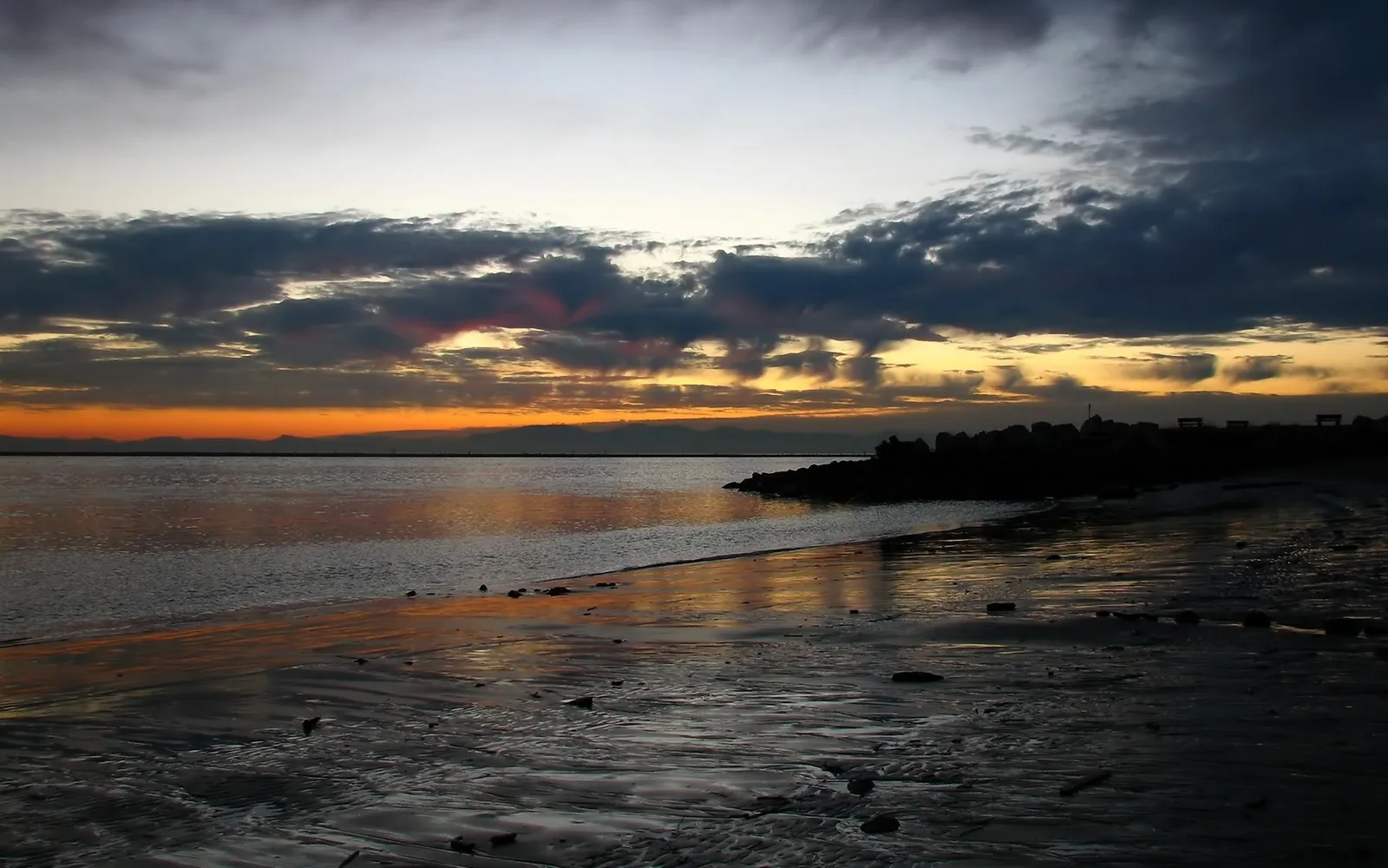 A dramatic sunset with dark clouds over a calm coastal waters and a rocky shoreline.