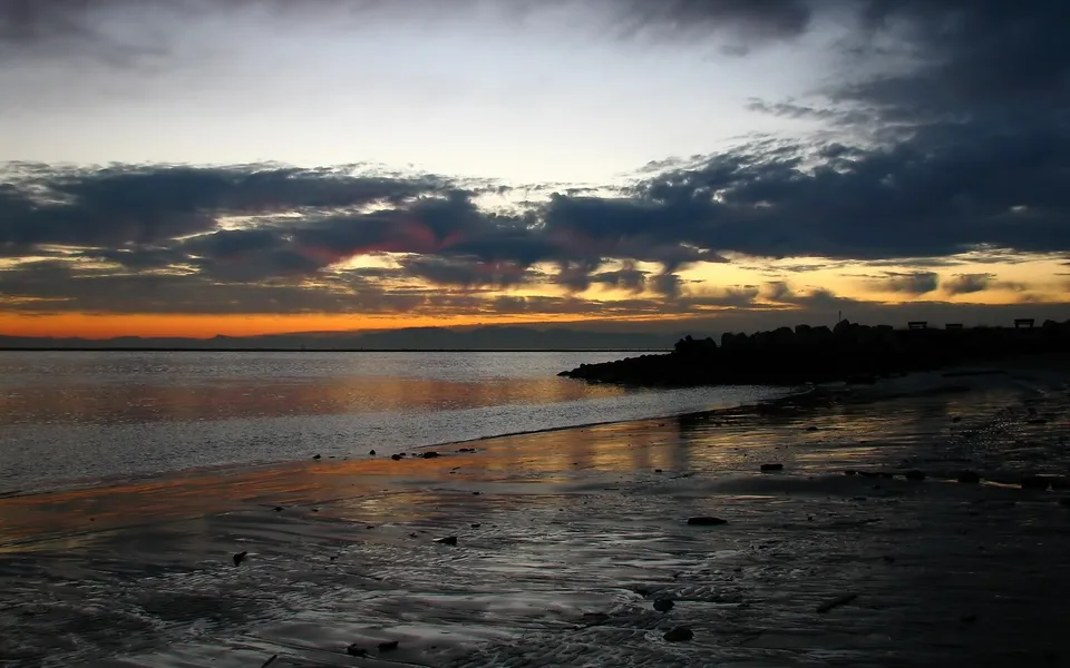 A dramatic sunset with dark clouds over a calm coastal waters and a rocky shoreline.