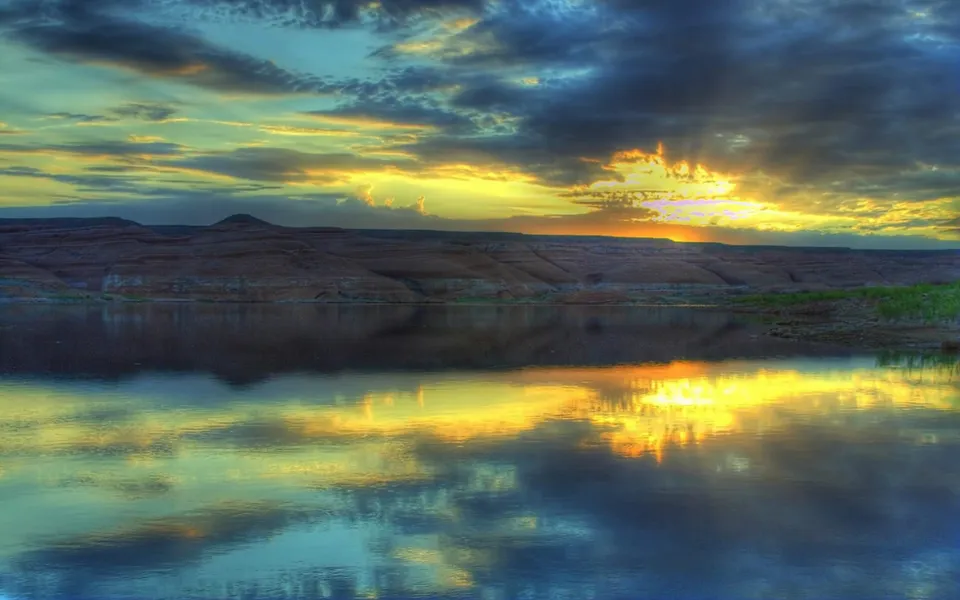 A vibrant sunset illuminates dramatic clouds over rugged hills and a reflective lake.