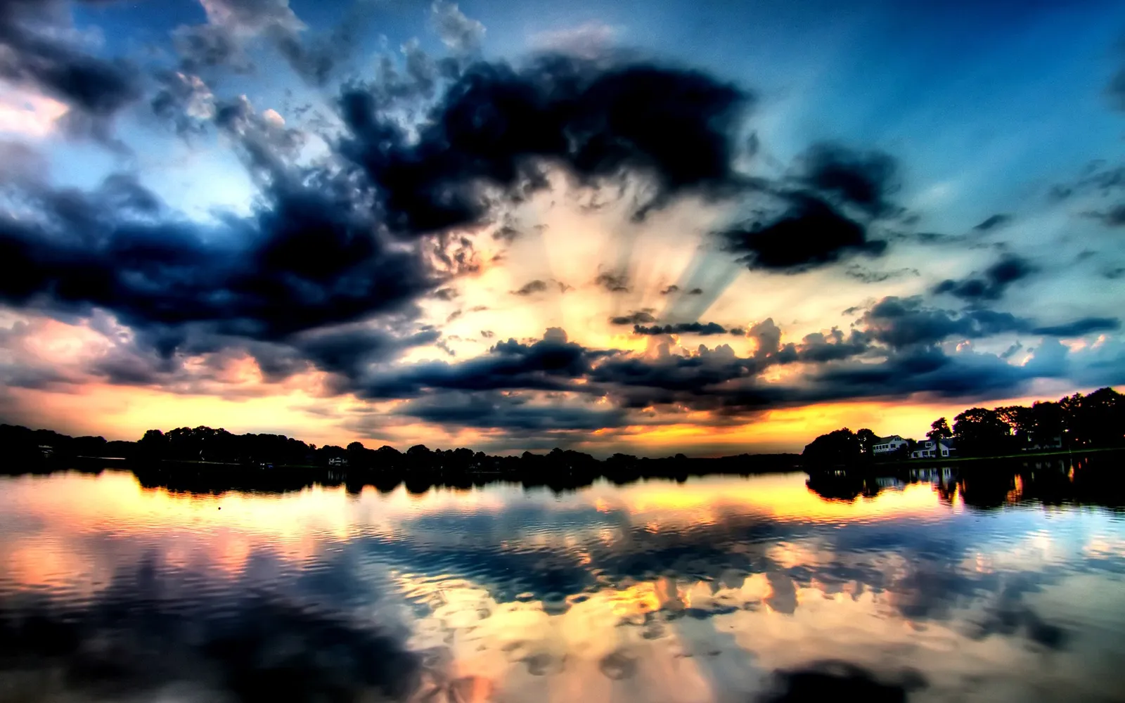A dramatic sunset with dark clouds and sunbeams reflecting on a calm lake.