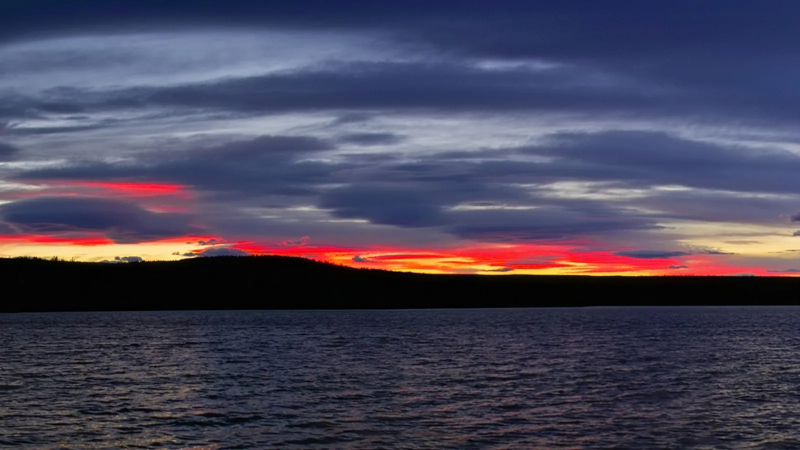 A dramatic sunset with vibrant red and orange hues near the horizon over a dark, calm lake.