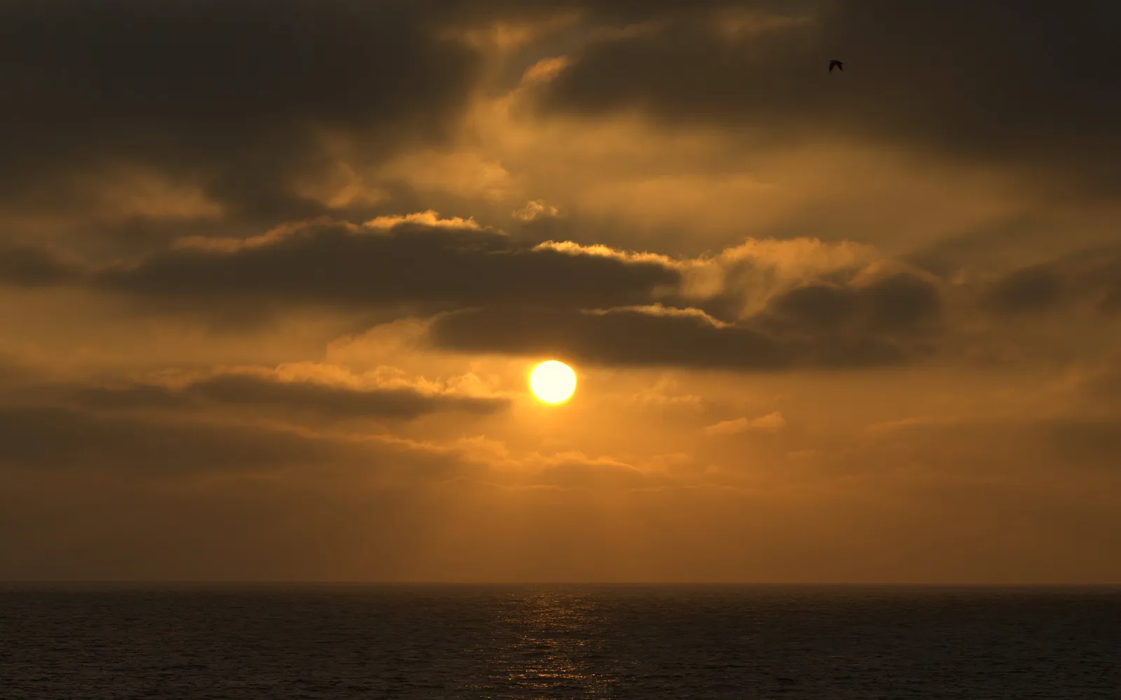 The sun is partially obscured by clouds as it sets over a dark, calm ocean.