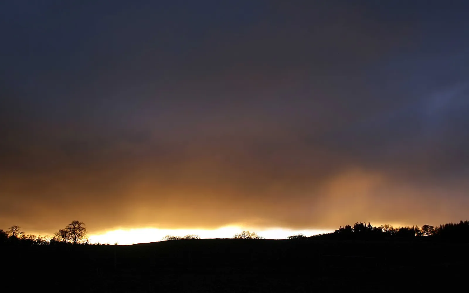 A dramatic sunset with a bright orange glow behind dark silhouetted hills and trees under a dark sky.