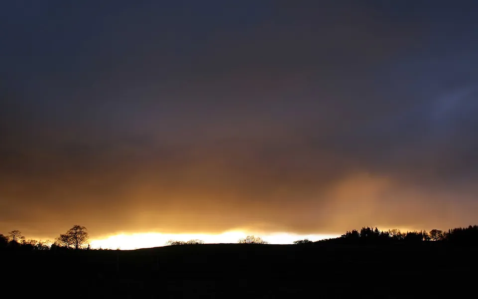 A dramatic sunset with a bright orange glow behind dark silhouetted hills and trees under a dark sky.