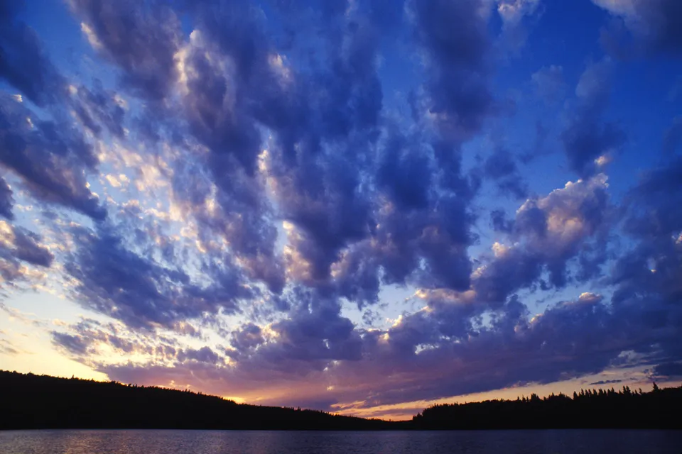 Dark clouds with illuminated edges against a deep blue and purple sky over a silhouetted treeline and water.