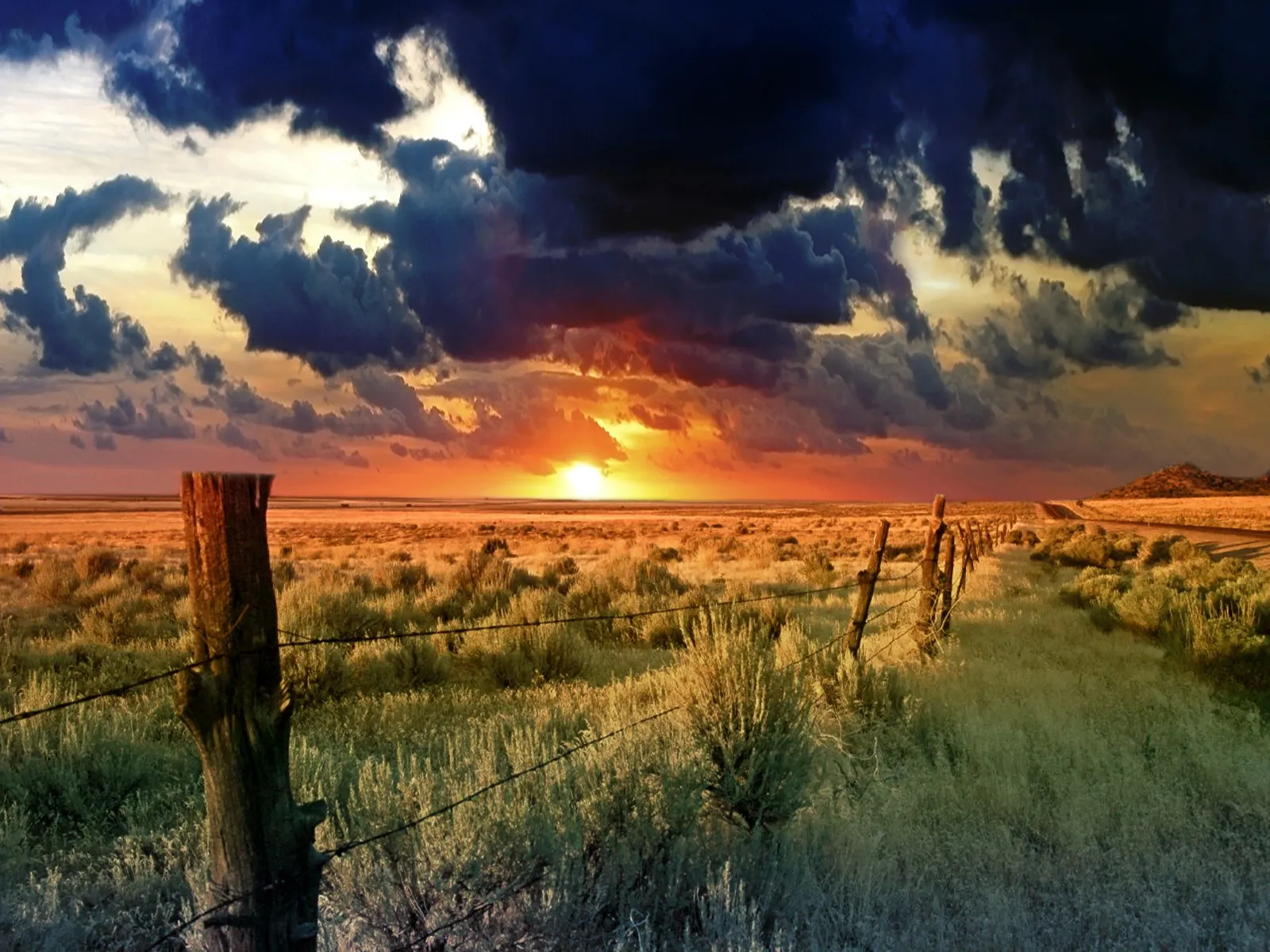 A dramatic sunset with dark clouds over a grassy prairie with a barbed wire fence in the foreground.