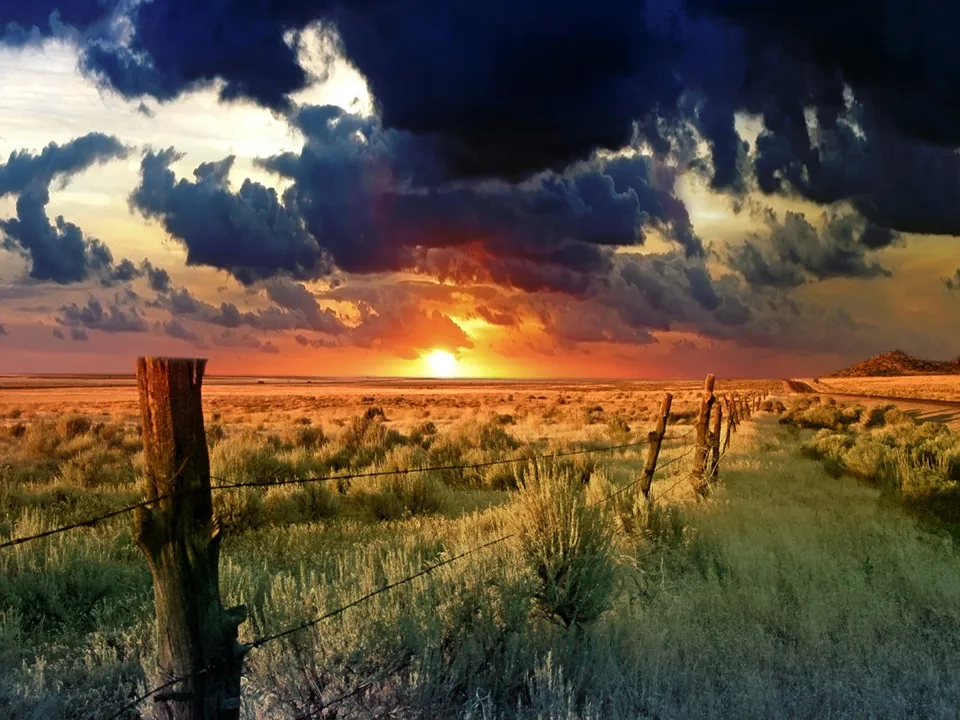 A dramatic sunset with dark clouds over a grassy prairie with a barbed wire fence in the foreground.