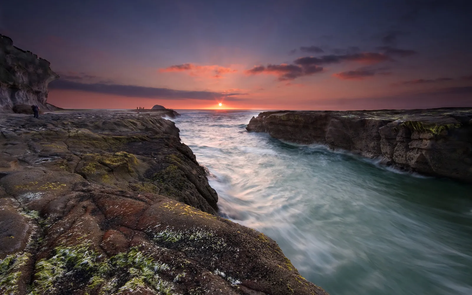 A sunset over a rocky shoreline with water flowing rapidly between rock formations.