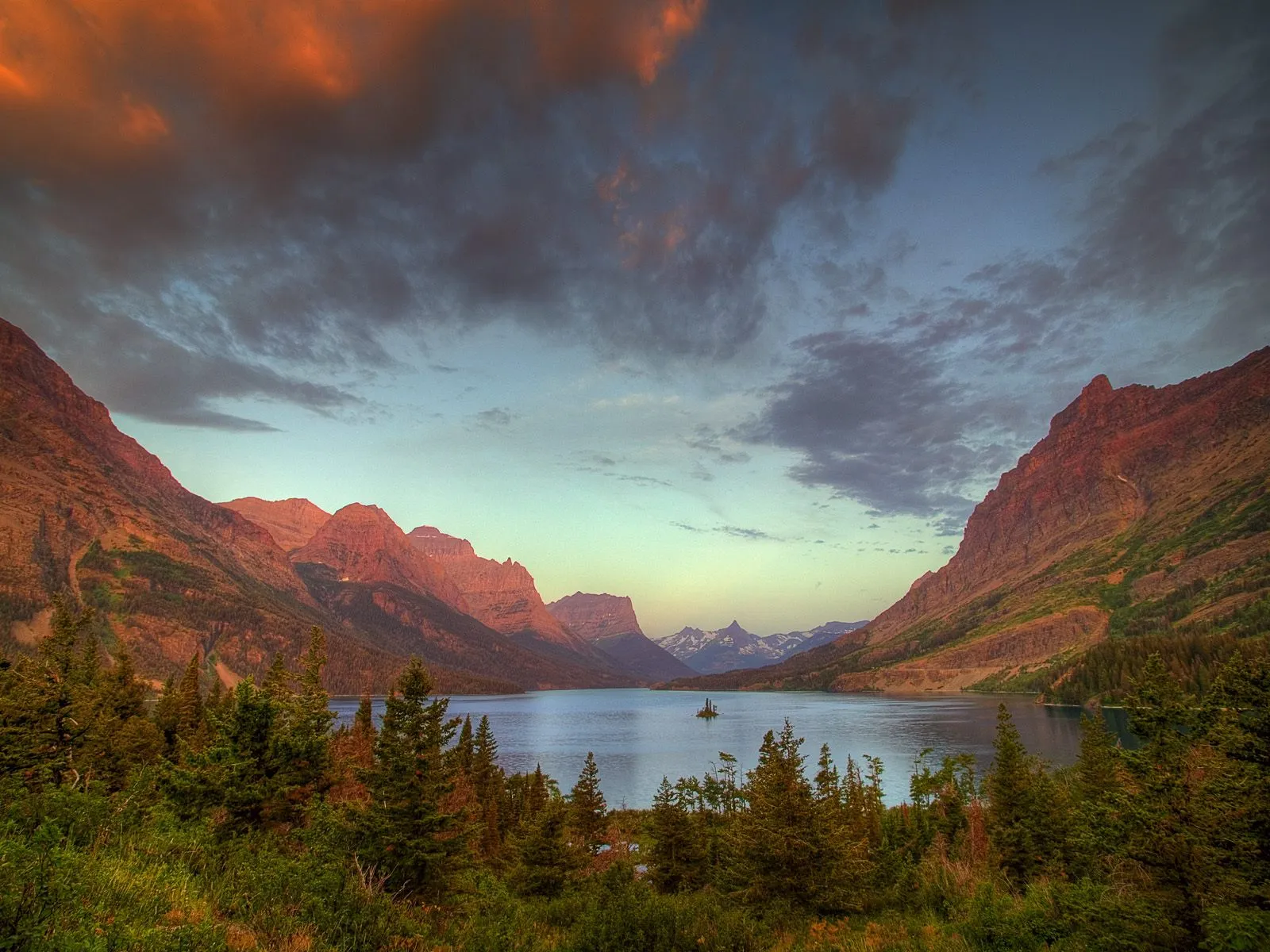 A serene lake reflects rugged mountains under a dramatic sunset sky.