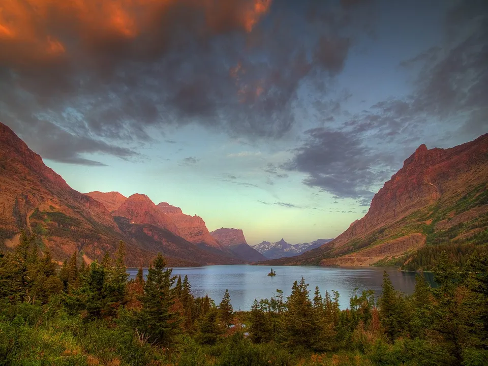 A serene lake reflects rugged mountains under a dramatic sunset sky.