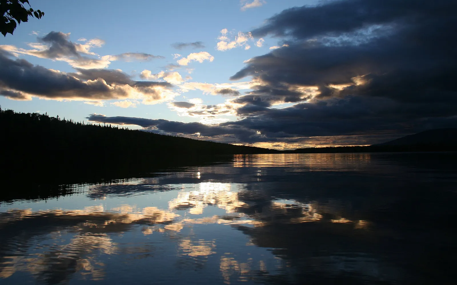 Setting sun illuminates dark clouds over a tranquil lake with a perfect water reflection.