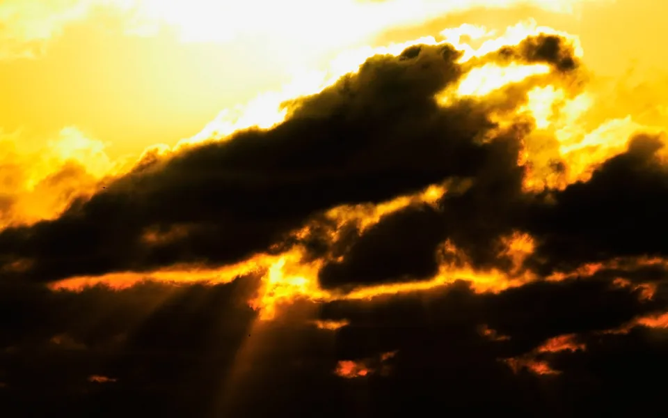 Dark clouds are illuminated by bright orange sunbeams breaking through during a dramatic sunset.