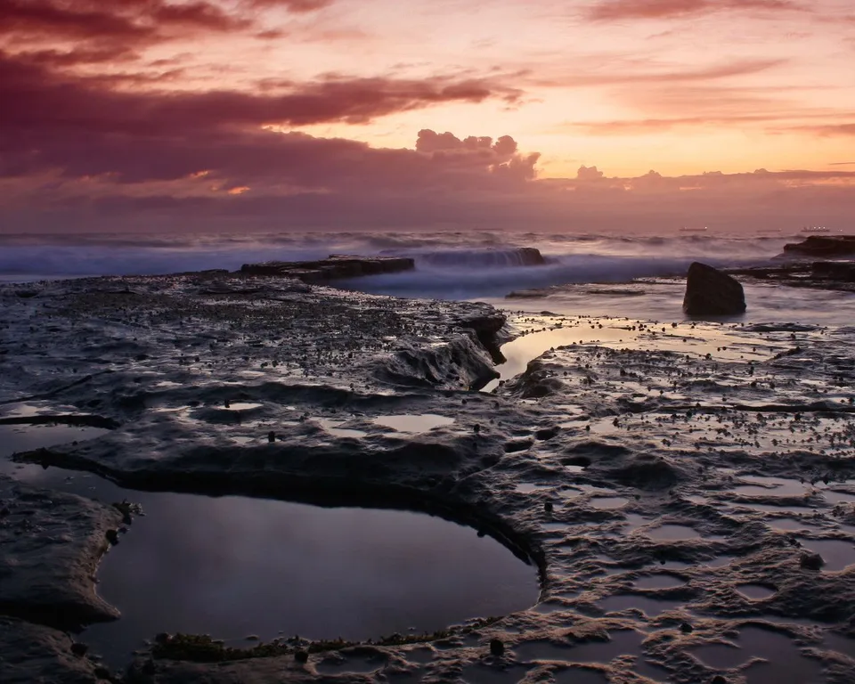 A dramatic sunset sky with pink, orange, and purple clouds over a wet, rocky shoreline with tide pools.
