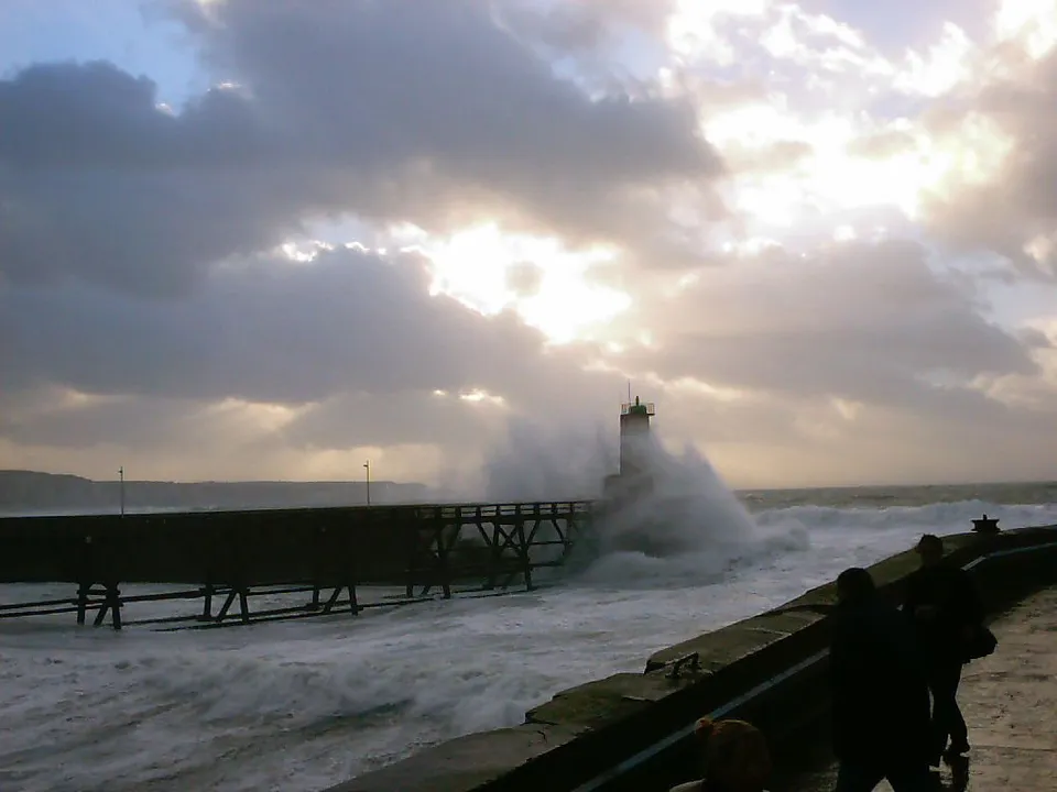 Dramatic waves surge against a wooden pier and lighthouse under dark, cloudy skies.