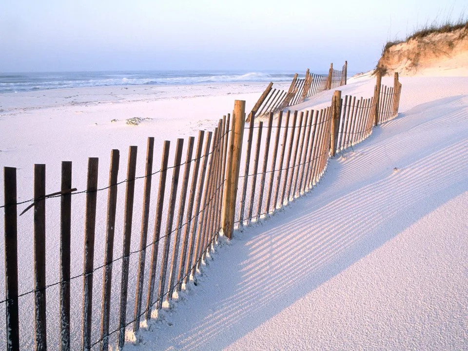 A wooden dune fence curves along a white sand beach with the ocean in the background.