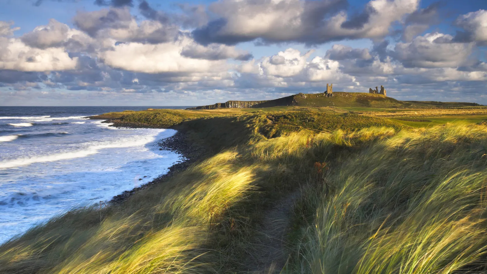 View of Dunstanburgh Castle ruins on a grassy cliff overlooking the sea, with waves crashing on the shore.