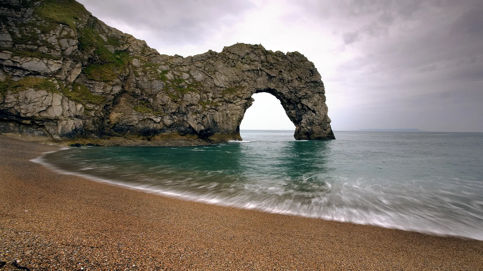 A natural limestone arch on a pebble beach with the sea flowing through it under a cloudy sky.