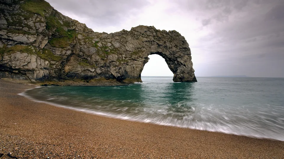 A natural limestone arch on a pebble beach with the sea flowing through it under a cloudy sky.