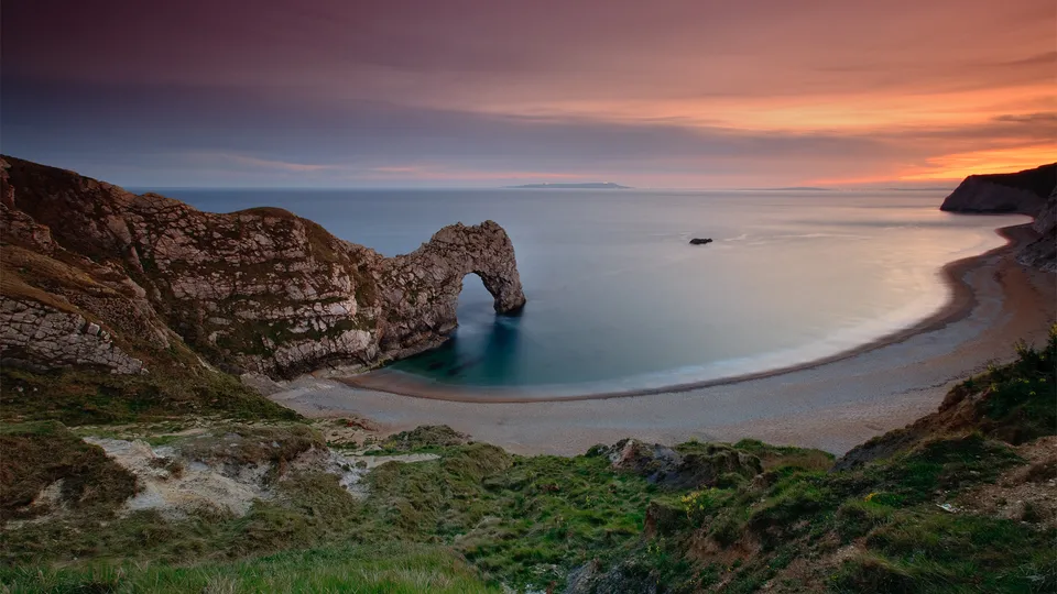 A natural limestone arch on a pebble beach at sunset with calm sea and colorful sky.