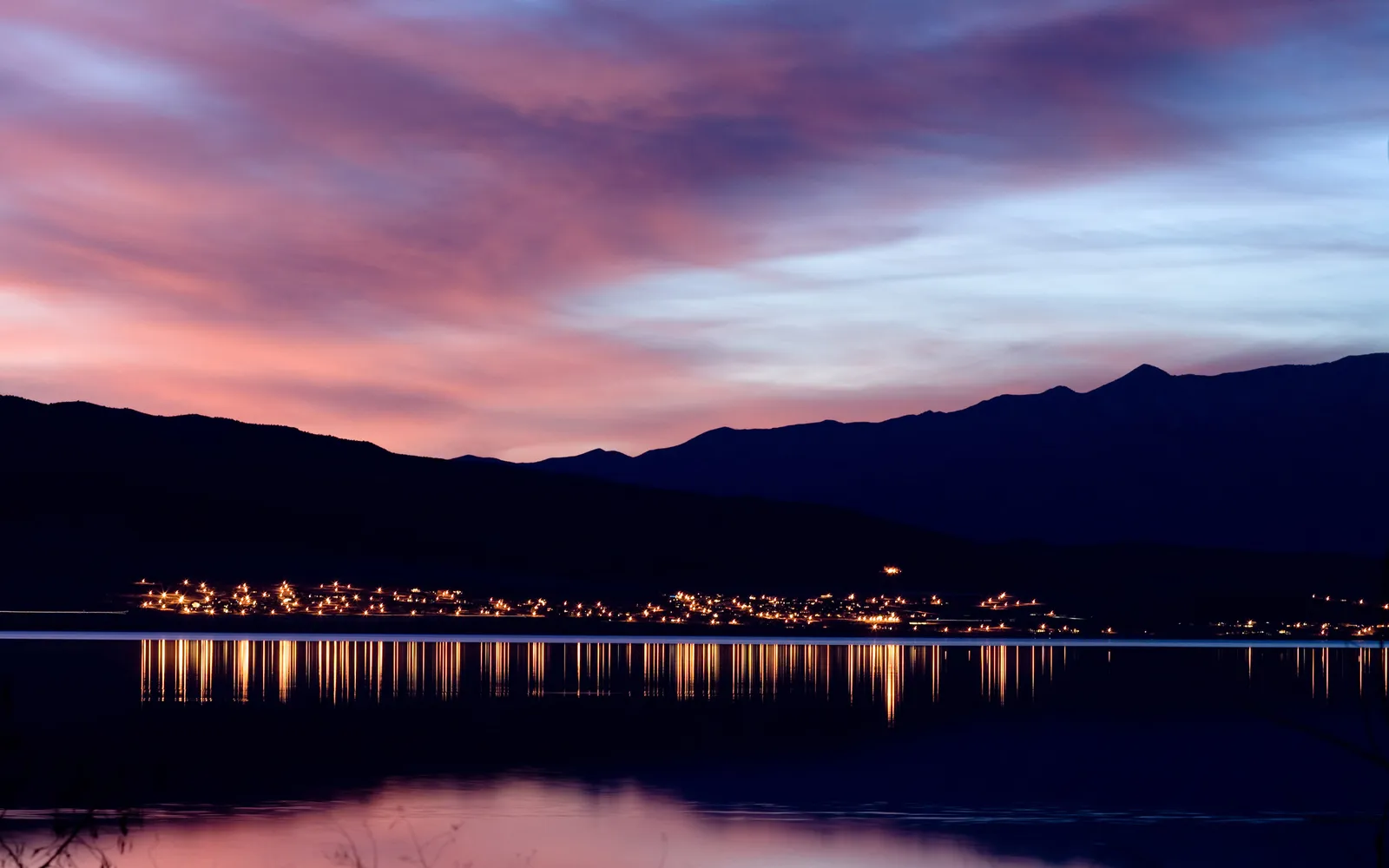 A town's lights reflect on a calm lake at dusk, with silhouetted mountains and colorful clouds in the sky.