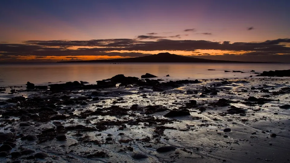 A dark, rocky shoreline in the foreground with the silhouette of Rangitoto Island in the distance under a purple and orange dusk sky.