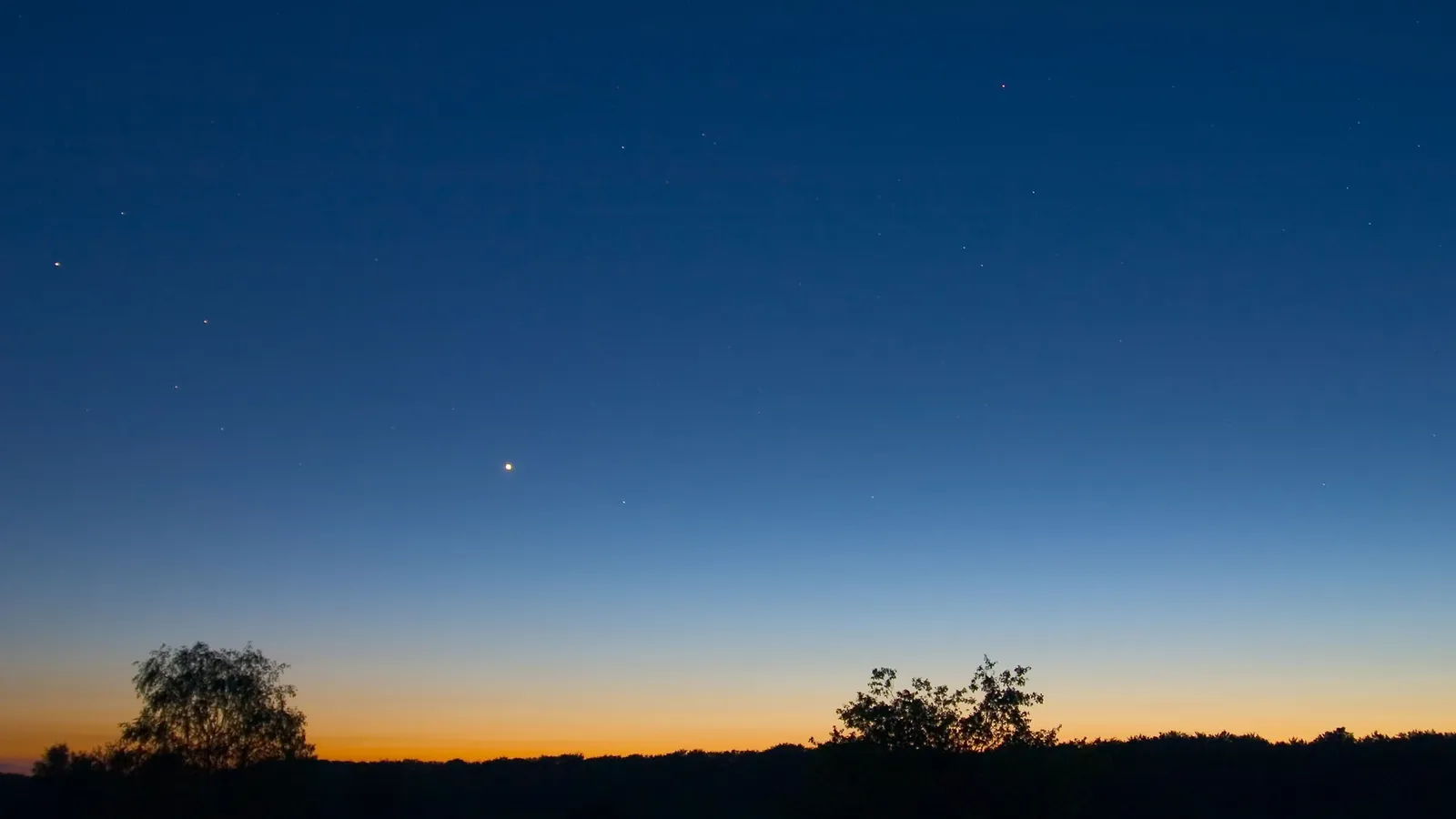 A gradient of deep blue to orange sky with scattered stars and a bright planet visible above a silhouetted treeline.