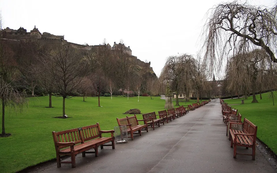 A view of Edinburgh Castle on a hill above a park with benches lining a pathway.