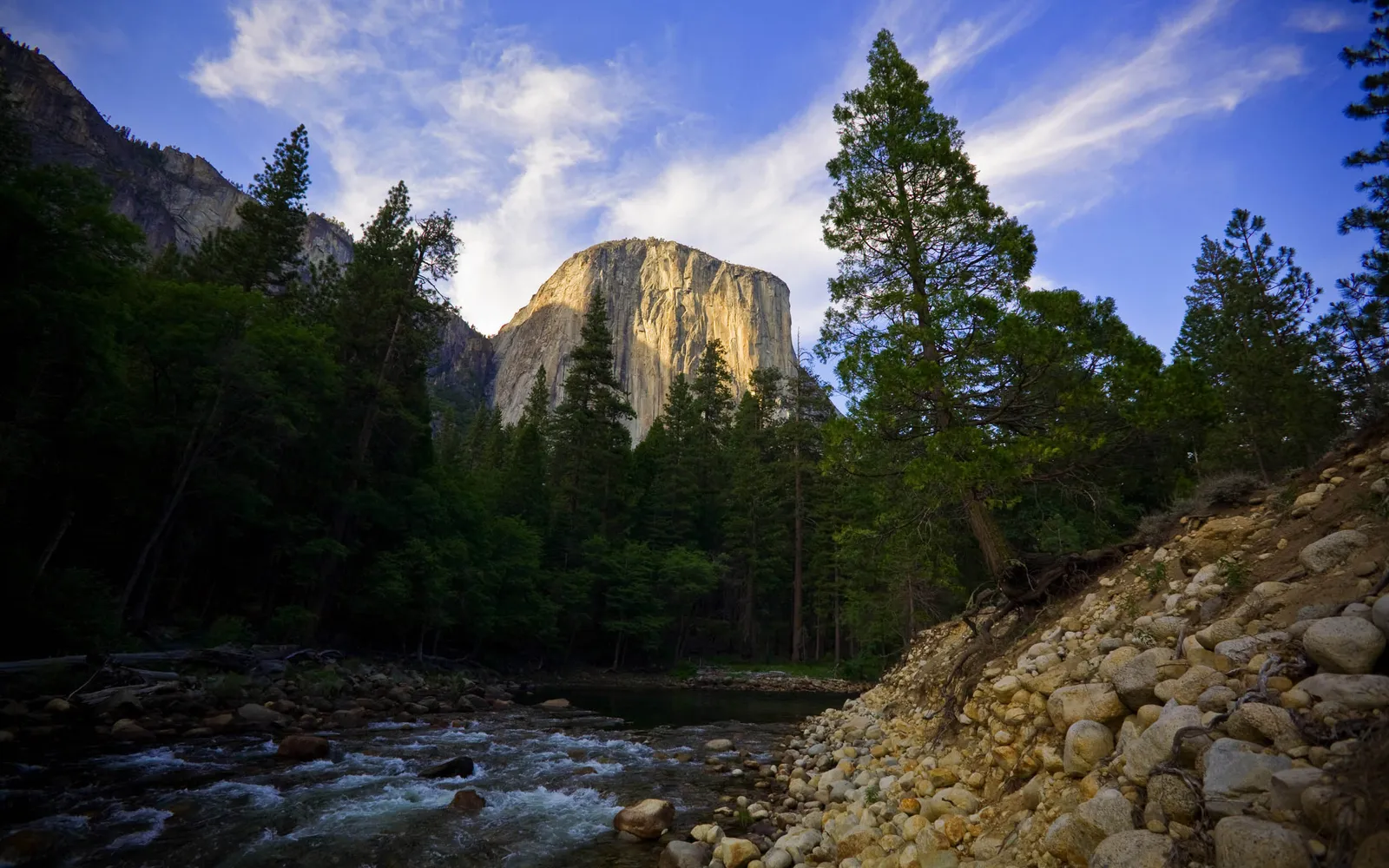 A majestic granite cliff face, El Capitan, is illuminated by the warm glow of the setting sun, with a river flowing in the foreground and pine trees surrounding it.