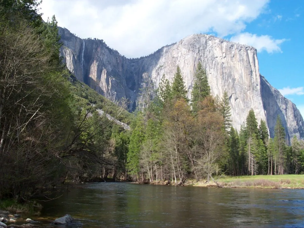 A wide, sheer granite cliff face, El Capitan, rises above a forested valley with a river in the foreground.