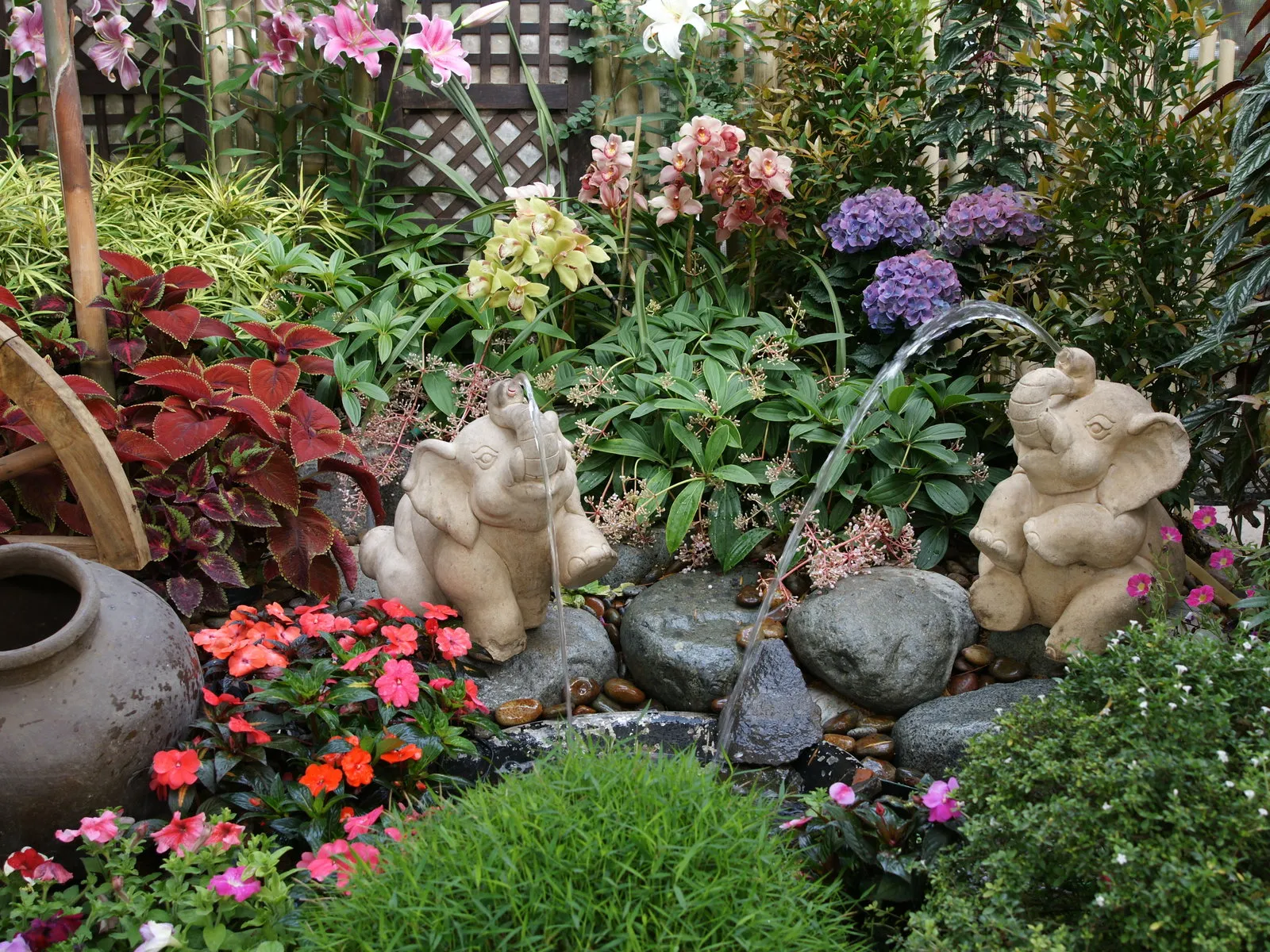 A garden fountain shaped like an elephant spouts water, surrounded by colorful flowers and lush greenery.