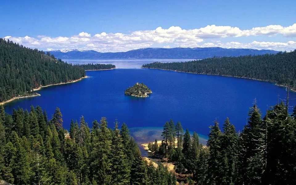 A vibrant blue lake with a small, tree-covered island, surrounded by dense pine forests and distant snow-capped mountains under a clear blue sky.