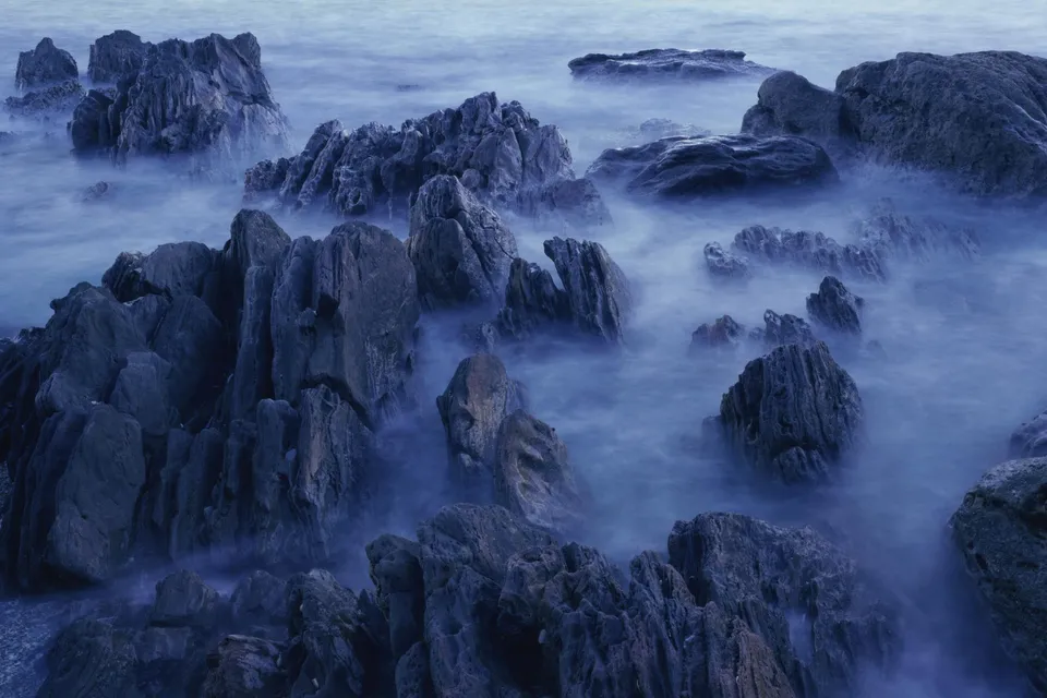 Jagged, dark rocks emerge from misty, blue water on a coastline.