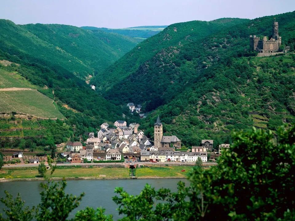 A scenic view of a European town with a river in the foreground and green hills surrounding it, with a castle on a hilltop.