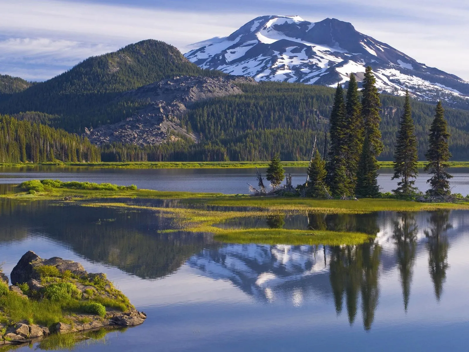 A snow-capped mountain and evergreen forest reflected in a calm lake under clear daylight.