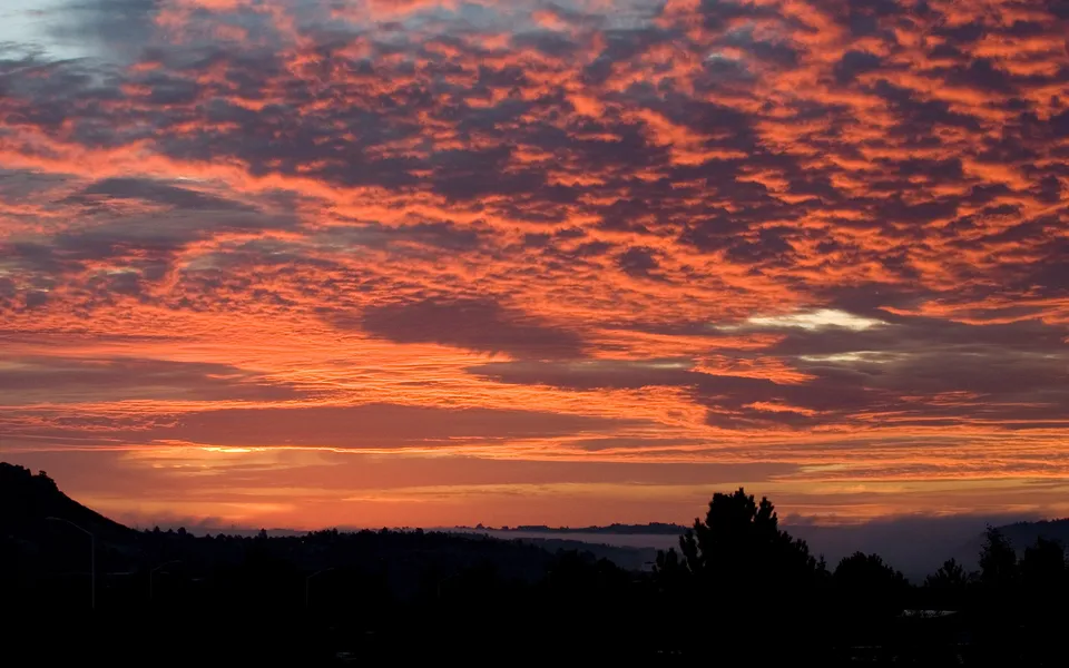 A dramatic sunset sky filled with fiery orange and red clouds above dark silhouetted hills and trees.